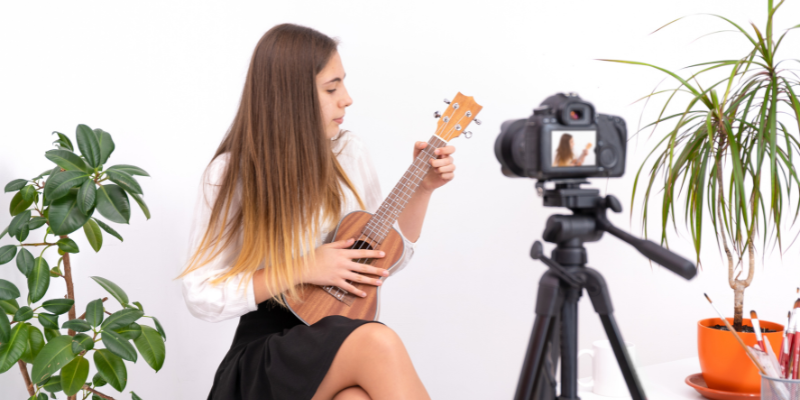 ragazza con capelli bruni seduta su uno sgabello che registra un video con la fotocamera mirrorless mentre suona l'ukulele